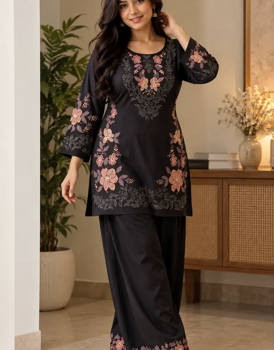 Woman wearing a black floral embroidered tunic with matching black palazzos and nude sandals in a warm indoor setting.